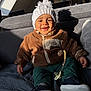 baby, child, smiling, hat, jacket, pacifier, indoor, couch, sunlight, shadow, cozy, warm_clothing, happy, seated, footwear, knit_hat, fleece_jacket, toy, person, home