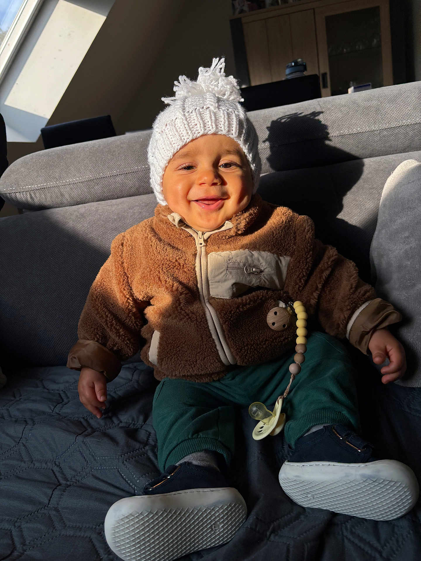 Ayden participe au concours pour gagner de l'argent avec cette photo : baby, child, smiling, hat, jacket, pacifier, indoor, couch, sunlight, shadow, cozy, warm_clothing, happy, seated, footwear, knit_hat, fleece_jacket, toy, person, home