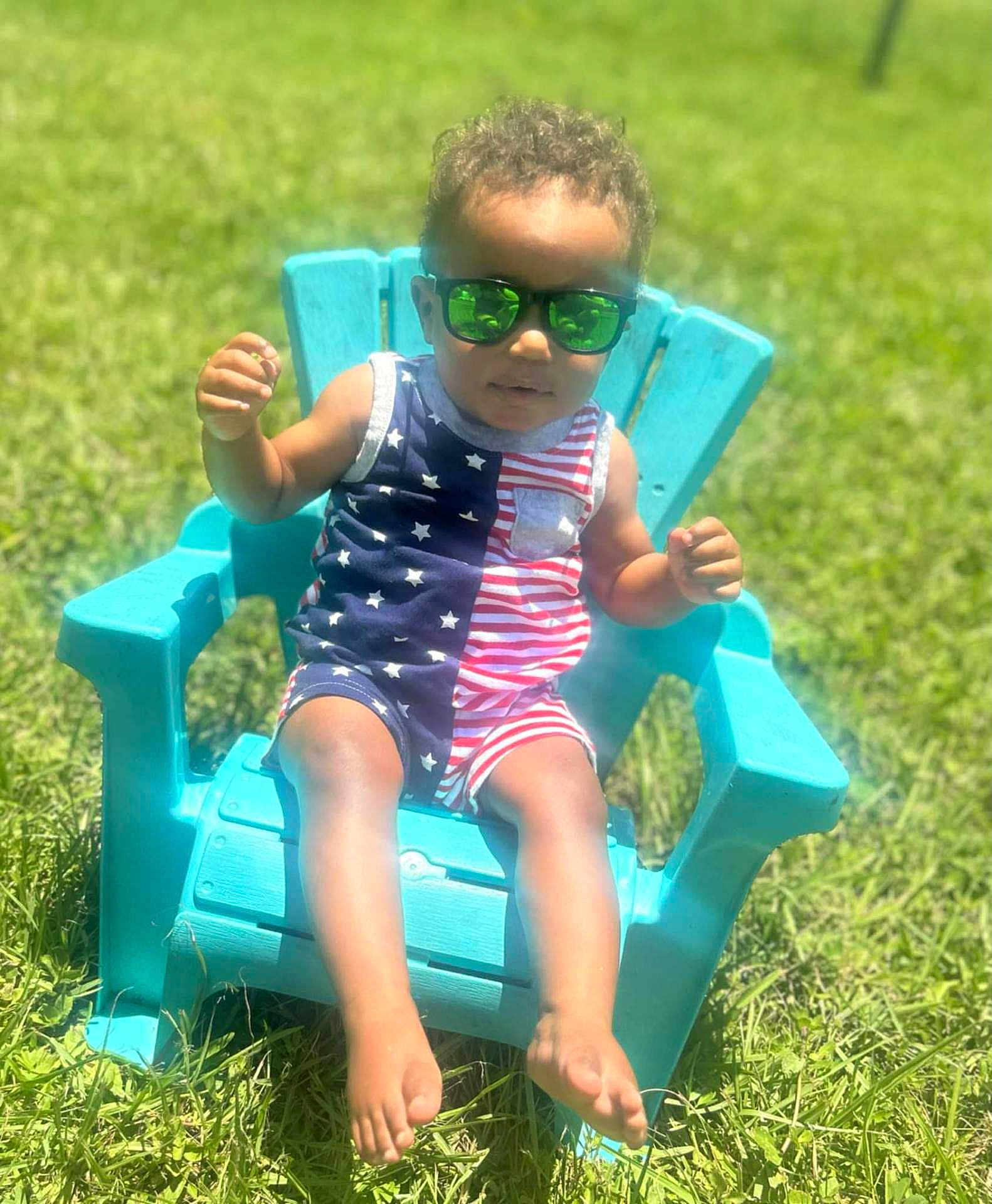 Omari is registered to the contest to win money with this photo: child, sunglasses, chair, outdoor, grass, sunny, blue, red, white, stars, stripes, summer, fashion, cute, person, feet, shorts, toddler, reflection, relaxing