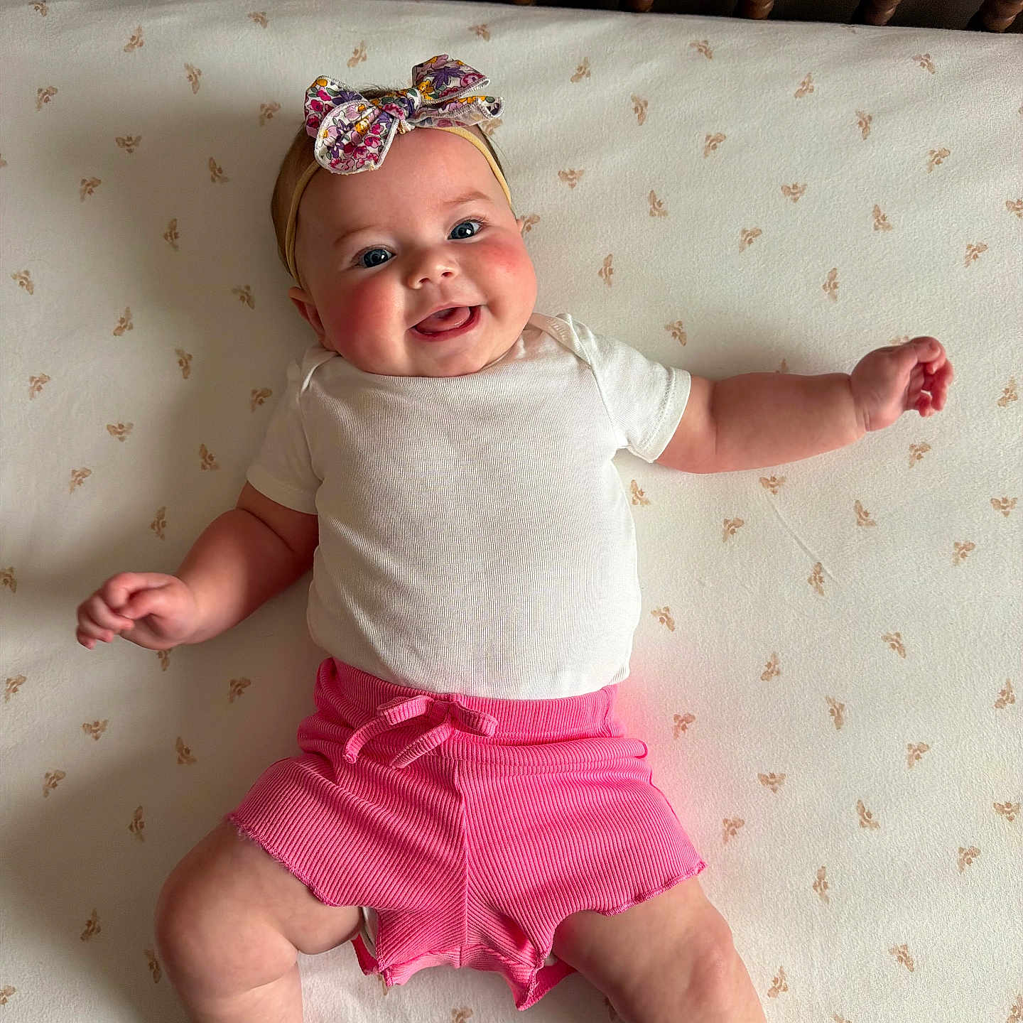 Landrie is registered to the contest to win money with this photo: baby, smiling, infant, headband, pink_shorts, white_shirt, lying_down, crib, mattress, pattern, happy, cute, chubby, skin, hand, foot, face, indoor, child, person
