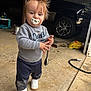 bag_of_material, child, concrete_floor, garage, grey_sweatshirt, hair_messy, hose, indoor, muddy_pants, pacifier, person, standing, toddler, tool, toy_in_background, truck, white_shoes, work_boots, wrench, young_child
