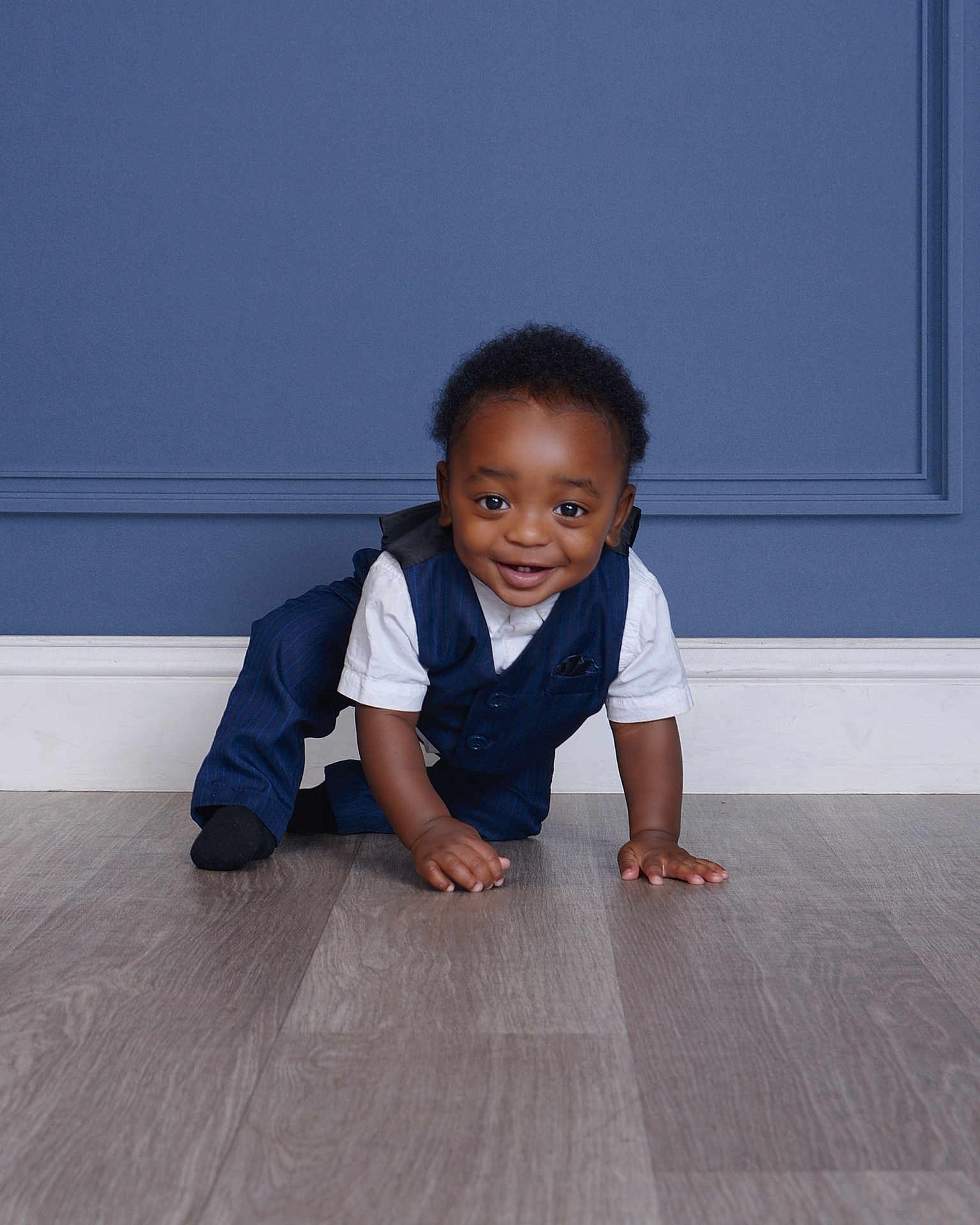 Hades is registered to the contest to win money with this photo: toddler, child, crawling, smiling, navy_suit, white_shirt, indoor, wooden_floor, blue_wall, paneled_wall, cute, happy, portrait, person, baby, fashion, formal_clothing, hands, feet, flooring