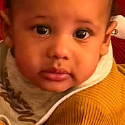 Hassun is registered to the contest to win money with this photo: baby, child, face, portrait, clothing, sweater, bib, red_background, close_up, young_child, indoor, curious, looking_at_camera, phone_screen, app_icons, notifications, mustard_color, soft_texture, warm_colors, casual