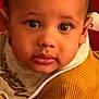 baby, child, face, portrait, clothing, sweater, bib, red_background, close_up, young_child, indoor, curious, looking_at_camera, phone_screen, app_icons, notifications, mustard_color, soft_texture, warm_colors, casual