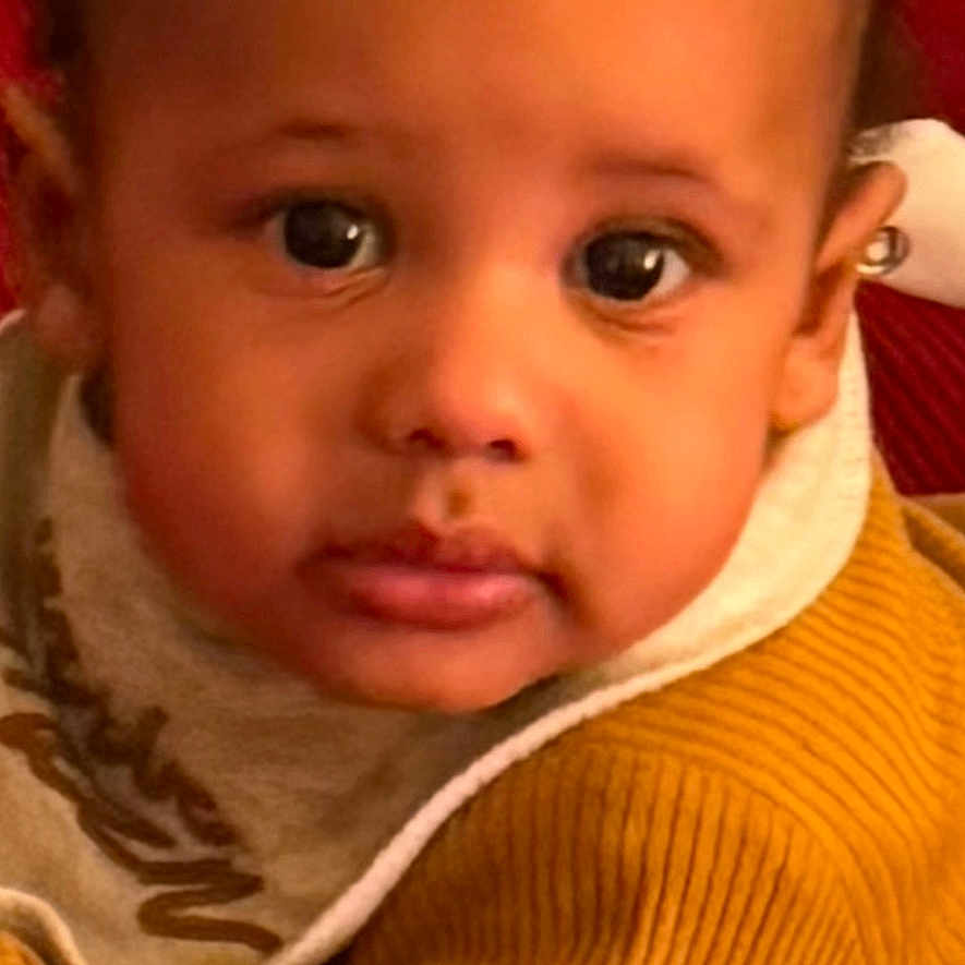 Hassun is registered to the contest to win money with this photo: app_icons, baby, bib, casual, child, close_up, clothing, curious, face, indoor, looking_at_camera, mustard_color, notifications, phone_screen, portrait, red_background, soft_texture, sweater, warm_colors, young_child