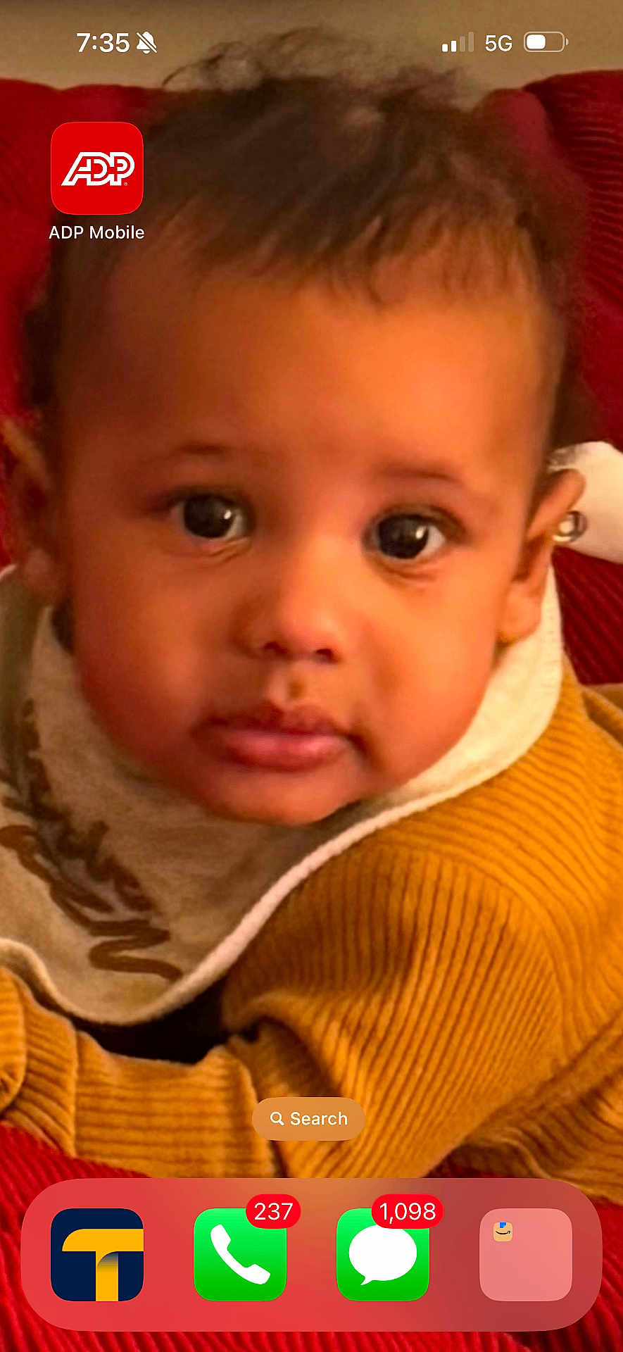 Hassun is registered to the contest to win money with this photo: baby, child, face, portrait, clothing, sweater, bib, red_background, close_up, young_child, indoor, curious, looking_at_camera, phone_screen, app_icons, notifications, mustard_color, soft_texture, warm_colors, casual