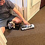 carpet, casual_clothing, cute, dog, door, floor, hand, happy, indoor, light, miniature, person, pet, playing, puppy, sitting, small_dog, toy_truck, wall, young_person