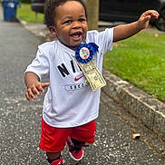 Zamir is registered to the contest to win money with this photo: toddler, child, smiling, happy, birthday, ribbon, dollar_bill, nike, soccer, red_shorts, sneakers, outdoor, street, sidewalk, greenery, car, celebration, walking, person, joyful
