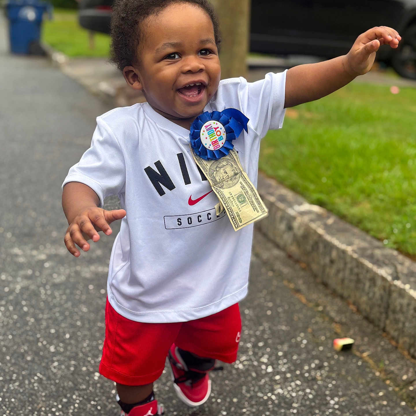 Zamir is registered to the contest to win money with this photo: toddler, child, smiling, happy, birthday, ribbon, dollar_bill, nike, soccer, red_shorts, sneakers, outdoor, street, sidewalk, greenery, car, celebration, walking, person, joyful