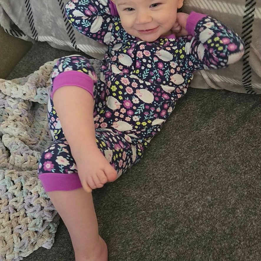 Melody joined the competition — help win amazing prizes! toddler, child, pajamas, smile, face, hair, ponytail, couch, blanket, carpet, floor, indoor, relaxed, playful, cute, person, clothing, patterned, happy, resting
