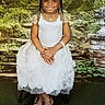 Skyla joined the competition — help win amazing prizes! child, girl, smile, dress, white_dress, sandals, bracelet, necklace, portrait, outdoor_background, garden, greenery, plants, stairs, stone_pillar, long_hair, braids, jewelry, happy, posing
