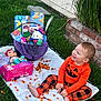 toddler, child, orange_clothing, pumpkin_face, candy_corn, blanket, grass, basket, toys, doll, snacks, brick_wall, outdoor, happy, smiling, holiday, fall, festive, playtime, cute