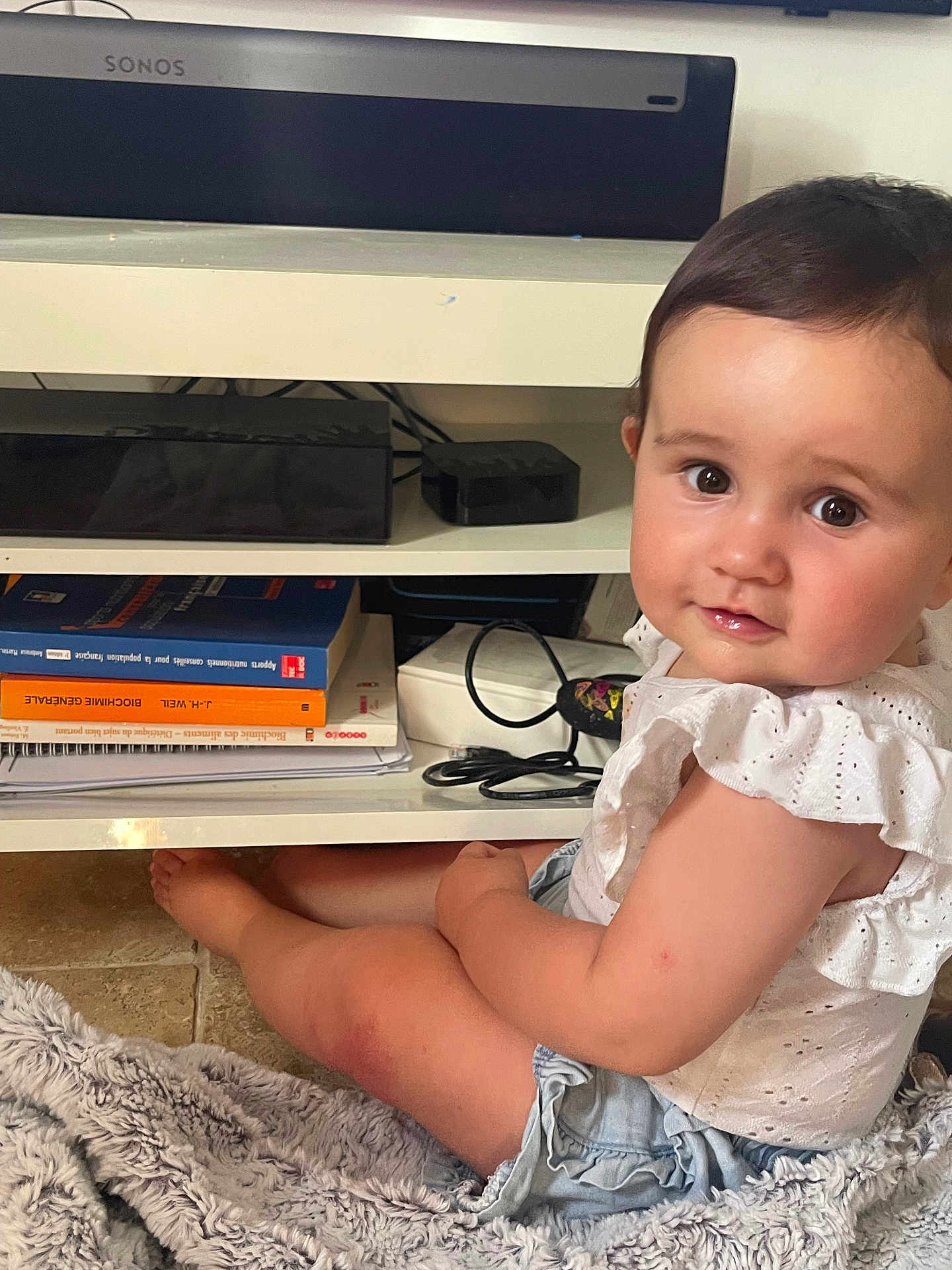 Nina a rejoint le concours — aidez-le/la à gagner de superbes lots ! baby, child, infant, sitting, white_top, denim_shorts, curly_hair, floor_rug, books, shelf, electronics, cables, indoor, portrait, looking_at_camera, cute, smiling, floor, home, furniture