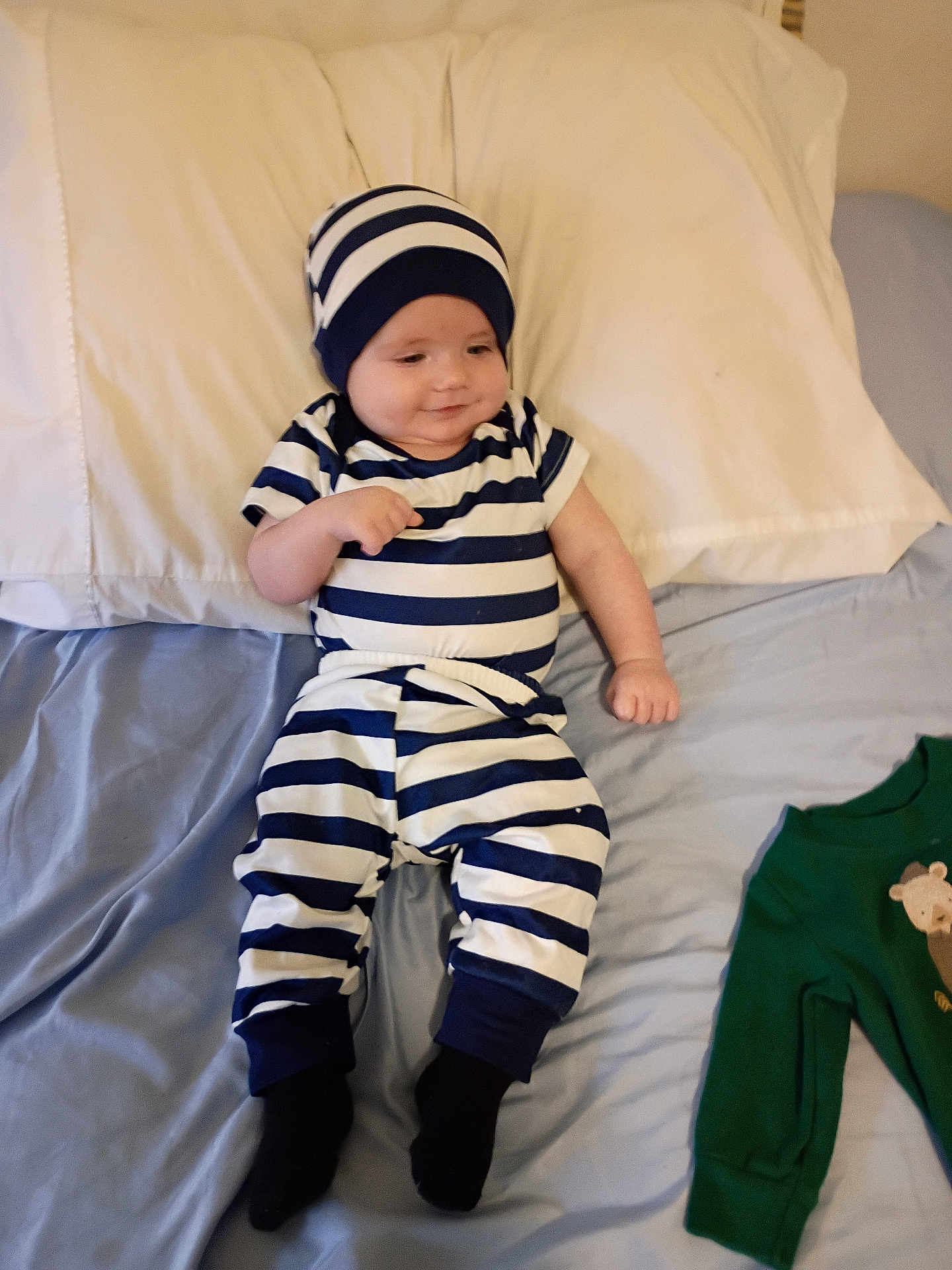 Stanley is registered to the contest to win money with this photo: baby, child, bed, pajamas, striped_clothing, hat, socks, pillow, blanket, bedroom, sleepwear, infant, cute, smiling, clothing, lying_down, green_onesie, soft_fabric, comfort, relaxed