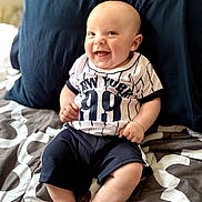 Amir is registered to the contest to win money with this photo: baby, smiling, sitting, bed, pillow, clothing, shorts, jersey, happy, infant, child, indoors, portrait, cute, person, skin, face, hands, feet, home