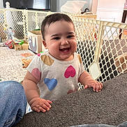 Ava joined the competition — help win amazing prizes! baby, child, smiling, indoor, living_room, couch, baby_gate, toys, colorful_clothing, heart_pattern, happy, person, furniture, carpet, television, jeans, hand, face, cute, home