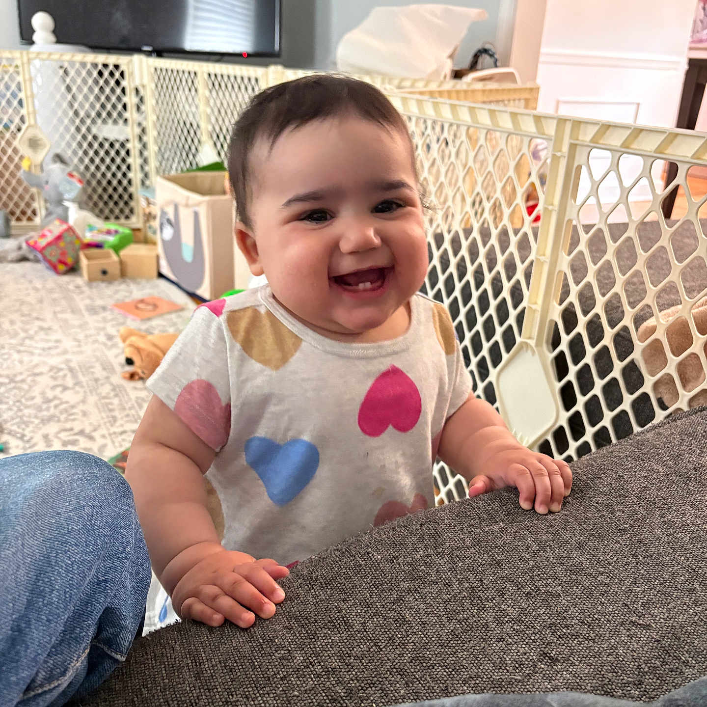 Ava joined the competition — help win amazing prizes! baby, child, smiling, indoor, living_room, couch, baby_gate, toys, colorful_clothing, heart_pattern, happy, person, furniture, carpet, television, jeans, hand, face, cute, home