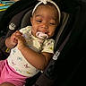baby, infant, car_seat, pacifier, headband, clothing, smile, happy, child, person, indoor, pink_shorts, white_shirt, butterfly_pattern, seatbelt, cute, young_child, seated, hands, face