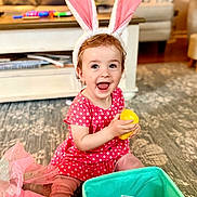 Isabelle is registered to the contest to win money with this photo: toddler, child, bunny_ears, pink_dress, yellow_egg, toy, carpet, living_room, smiling, happy, sitting, playtime, indoors, cute, play, colorful, basket, furniture, home, portrait