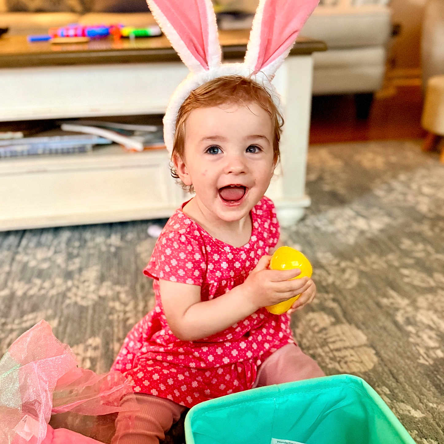 Isabelle is registered to the contest to win money with this photo: toddler, child, bunny_ears, pink_dress, yellow_egg, toy, carpet, living_room, smiling, happy, sitting, playtime, indoors, cute, play, colorful, basket, furniture, home, portrait