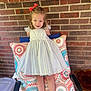 child, girl, dress, white_dress, bow, red_bow, cushion, patterned_cushion, brick_wall, smiling, happy, portrait, outdoor, chair, legs, barefoot, cute, young, person, playful