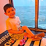 child, boy, smiling, sitting, window, sea, water, toy, rubber_duck, sandals, shorts, tshirt, indoor, playtime, happy, casual_clothing, legs_crossed, daylight, reflection, relaxation