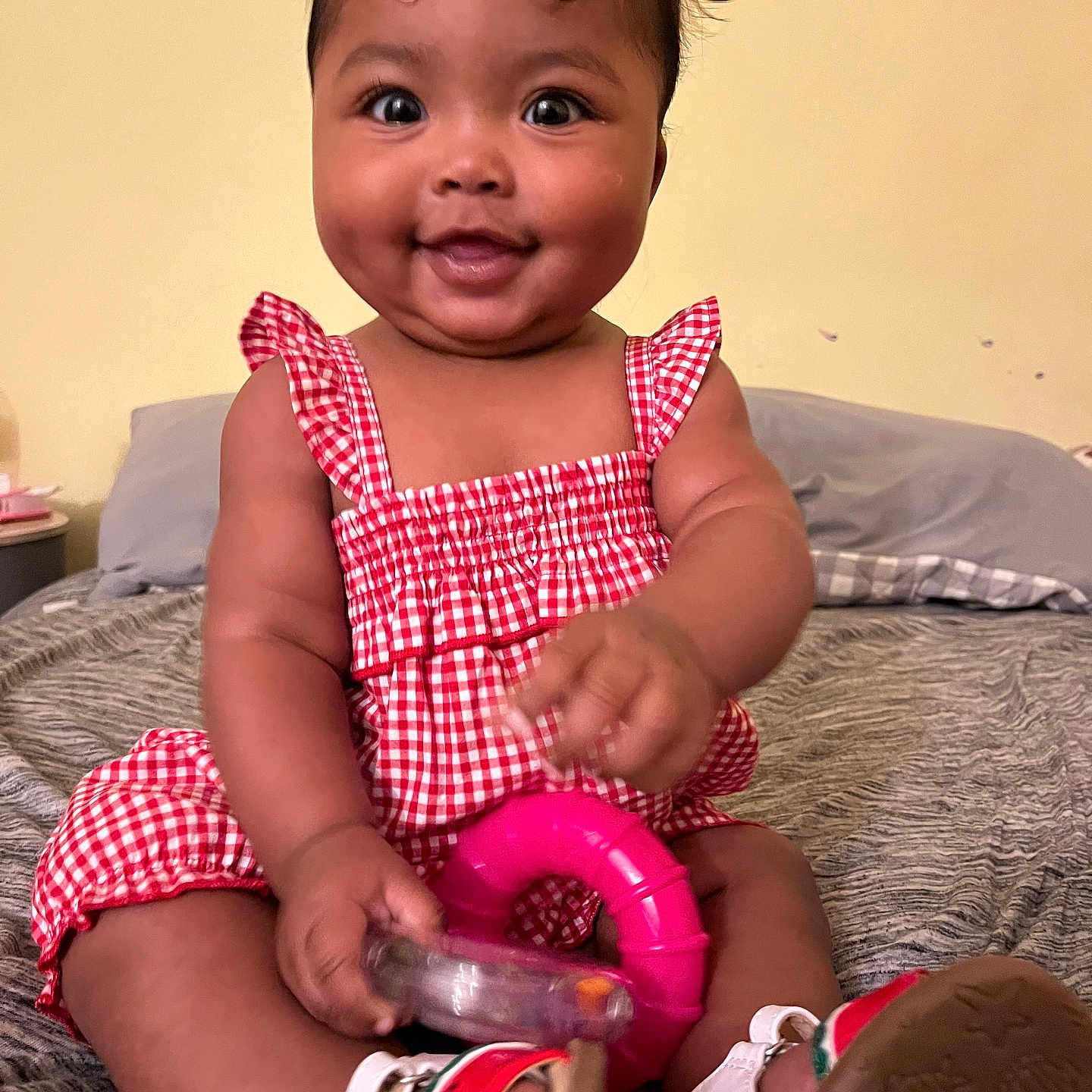 Amaia joined the competition — help win amazing prizes! baby, child, smiling, sitting, red_dress, checkered, toy, pink_toy, bed, sandals, footwear, indoor, cute, happy, dimples, person, infant, clothing, hand, face