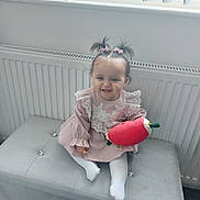 Isabella is registered to the contest to win money with this photo: baby, child, girl, smiling, pink_dress, lace, pony_tail, hair_bows, plush_toy, red_toy, bench, cushion, window, curtains, radiator, white_tights, indoor, happy, cute, portrait