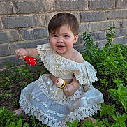 Zia is registered to the contest to win money with this photo: baby, child, girl, dress, maracas, plants, greenery, brick_wall, smiling, happy, outdoor, nature, sitting, lace, toy, cute, playful, young, person, portrait