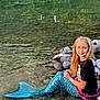 child, girl, mermaid_tail, water, river, rocks, smile, long_hair, nature, outdoor, reflection, blue, black_tshirt, playful, sitting, shallow_water, daylight, scenery, casual_clothing, happy