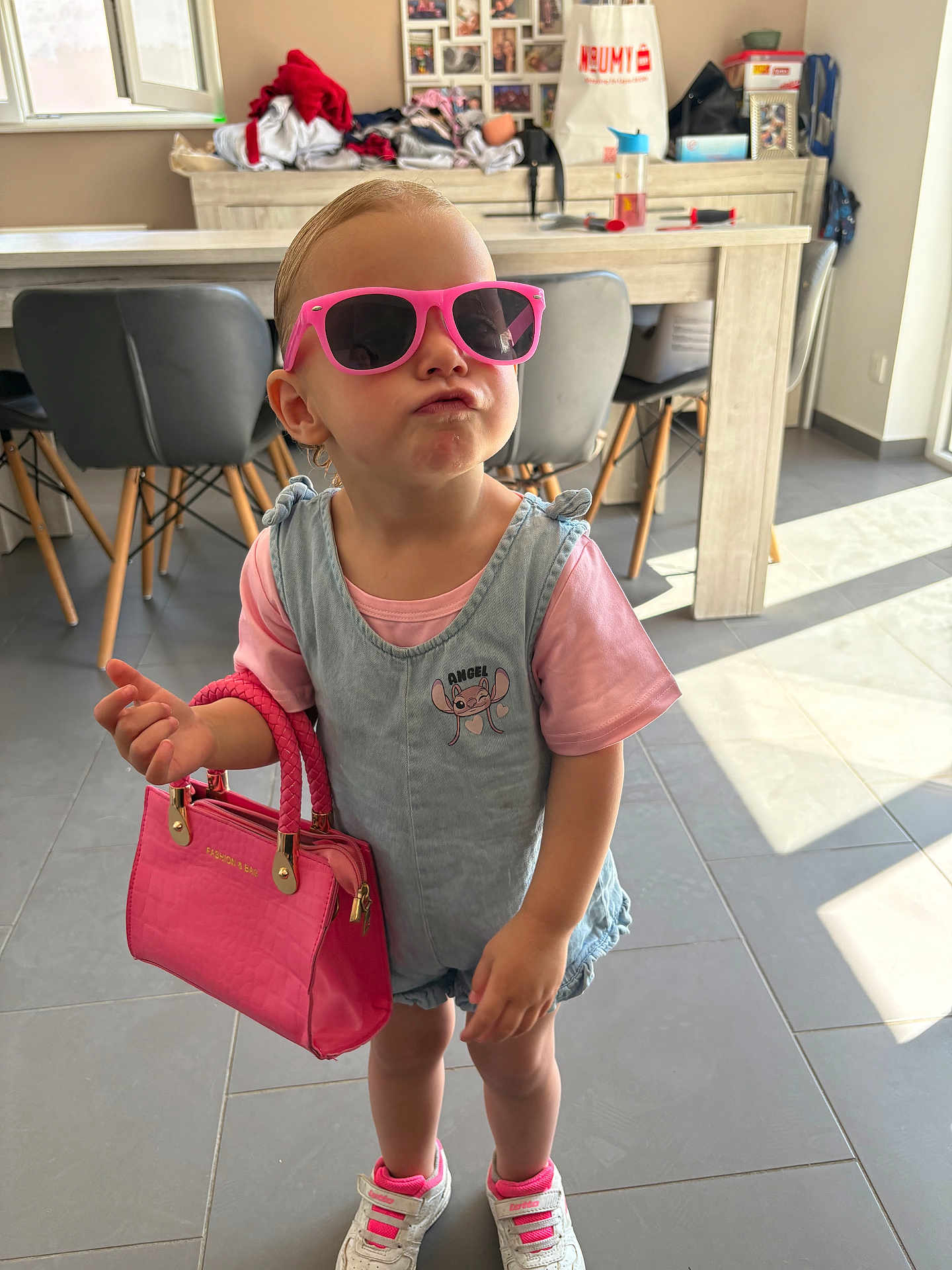 Maëlya participe au concours pour gagner de l'argent avec cette photo : attitude, chairs, child, cute, denim_overall, fashion, floor_tiles, handbag, indoor, kitchen, modern, person, pink_shirt, pink_sunglasses, playful, pout, sneakers, sunlight, table, toddler