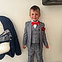 boy, casual, chair, child, clothing, face, floor, formal_wear, grey_suit, indoor, jacket, pants, person, portrait, red_bow_tie, red_flower, smiling, standing, suit, white_wall
