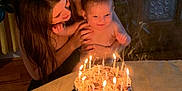 Levi is registered to the contest to win money with this photo: baby, woman, birthday_cake, candles, indoor, celebration, table, smiling, happy, person, cake, light, warm, curtains, cabinet, plant, home, family, together, joy