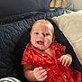 Levi is registered to the contest to win money with this photo: baby, smiling, red_shirt, quilt, cushion, indoor, happy, child, portrait, infant, face, person, cute, relaxed, seated, home, young, hands, expressive, joyful
