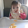 Cheyanne is registered to the contest to win money with this photo: bib, cheese_puffs, child, curious, eating, face, food, hair, hand, high_chair, indoor, natural_light, person, plastic_tray, red_clothing, seat, snack, table, toddler, window
