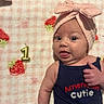 baby, infant, cute, headband, bow, tongue_out, birthday_candle, number_one, blanket, strawberry_pattern, blue_tank_top, text_on_clothing, lying_down, playful, portrait, child, skin, face, expression, celebration