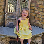 Wynter is registered to the contest to win money with this photo: child, girl, yellow_dress, blue_crocs, golf_putter, bench, stone_bench, brick_wall, wooden_door, outdoor, smiling, happy, portrait, sitting, toy, playful, sunlight, cute, young_child, fashion