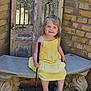 child, girl, yellow_dress, blue_crocs, golf_putter, bench, stone_bench, brick_wall, wooden_door, outdoor, smiling, happy, portrait, sitting, toy, playful, sunlight, cute, young_child, fashion