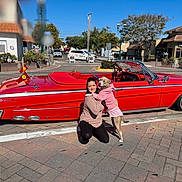 Hunnybun is registered to the contest to win money with this photo: woman, dog, red_car, vintage_car, convertible, sidewalk, street, tree, building, blue_sky, sunny_day, outdoor, kneeling, pet, clothing, hoodie, hugging, happy, urban, transportation