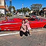 woman, dog, red_car, vintage_car, convertible, sidewalk, street, tree, building, blue_sky, sunny_day, outdoor, kneeling, pet, clothing, hoodie, hugging, happy, urban, transportation