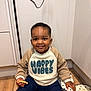 child, toddler, smiling, sweatshirt, happy_vibes, cookie, kitchen, floor, wooden_floor, oven_mitt, heart_pattern, indoor, person, casual_clothing, happy, cute, sitting, young_child, shadow, candid