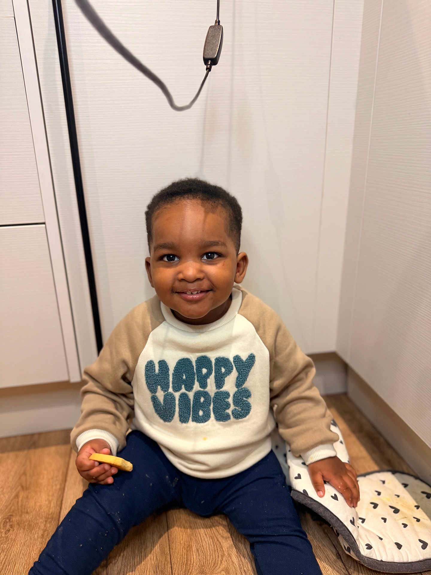 Bryson joined the competition — help win amazing prizes! child, toddler, smiling, sweatshirt, happy_vibes, cookie, kitchen, floor, wooden_floor, oven_mitt, heart_pattern, indoor, person, casual_clothing, happy, cute, sitting, young_child, shadow, candid