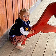 Yanaëlle participe au concours pour gagner de l'argent avec cette photo : child, toddler, smiling, wooden_wall, wooden_floor, red_bench, bench, blue_eyes, sweater, dress, hands, shoes, sitting, portrait, planks, outdoors, wood_paneling, cute, light, young_child