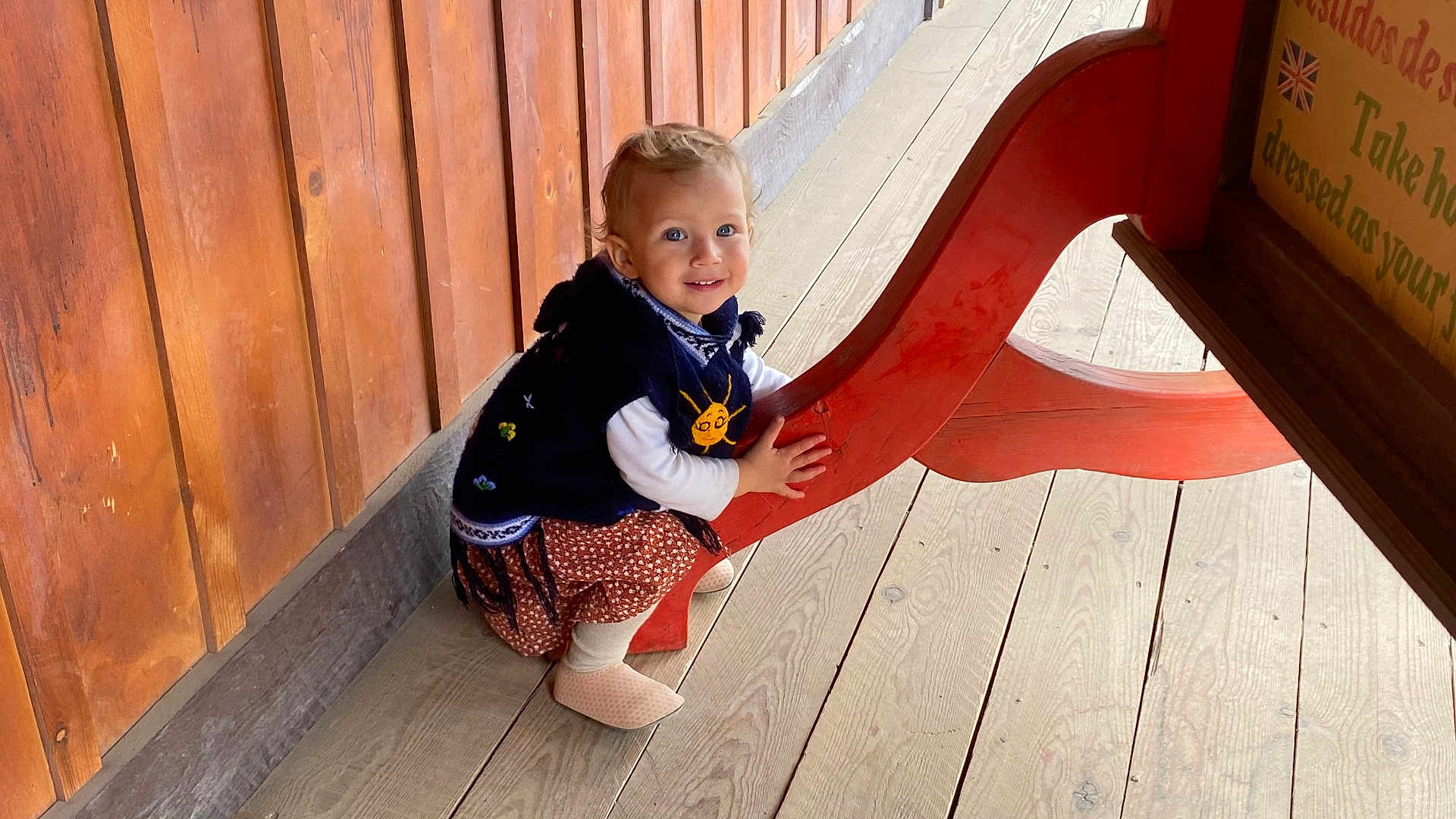 Yanaëlle participe au concours pour gagner de l'argent avec cette photo : child, toddler, smiling, wooden_wall, wooden_floor, red_bench, bench, blue_eyes, sweater, dress, hands, shoes, sitting, portrait, planks, outdoors, wood_paneling, cute, light, young_child