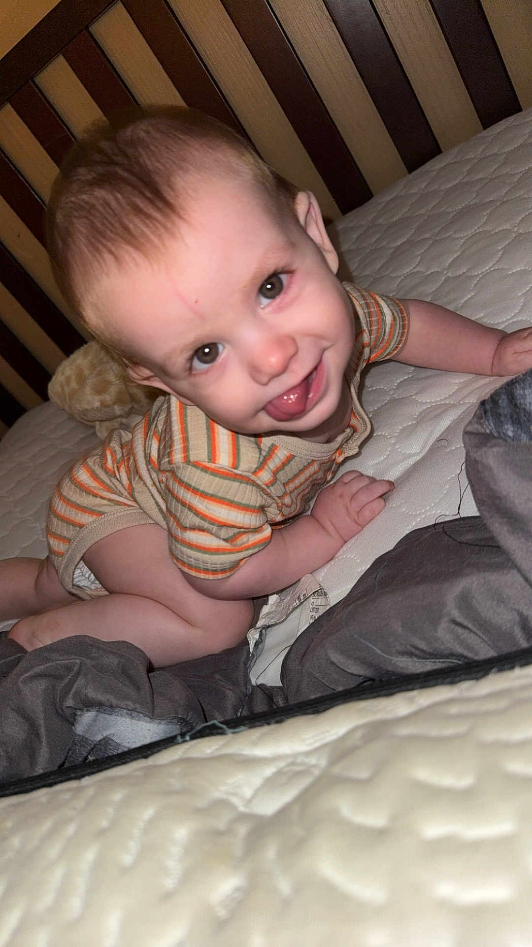 Damon is registered to the contest to win money with this photo: baby, child, infant, bed, crib, striped_clothing, tongue_out, smiling, crawling, cute, indoor, mischievous, wooden_crib, mattress, bedding, pale_skin, short_hair, playful, happy, young_child
