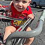 toddler, child, shopping_cart, red_clothing, outdoor, parking_lot, sunlight, barefoot, metal_bars, toy_truck_graphic, smiling, face, legs, hand, trees, vehicle, asphalt, daytime, person, holding
