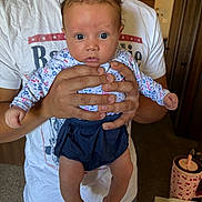 Georgia is registered to the contest to win money with this photo: baby, infant, person, hands, clothing, indoor, blue_eyes, shorts, long_sleeve, tshirt, adult, carpet, door, furniture, cup, table, curious, holding, portrait, closeup