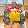 Jamila joined the competition — help win amazing prizes! baby, child, plush_car, costume, indoor, person, legs, shorts, wooden_chair, window, natural_light, happy, smiling, toy, floor, fabric, seat, hand, feet, face