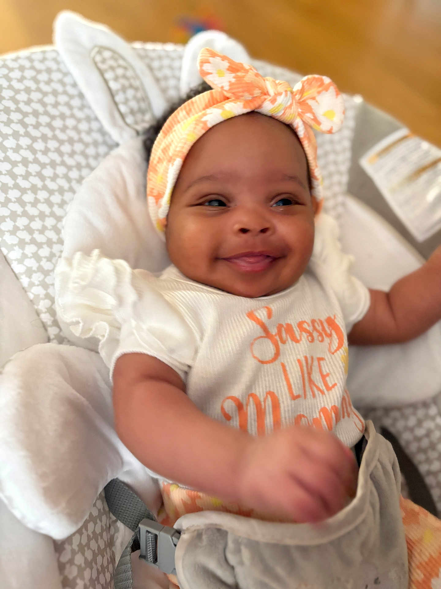 Jamila is registered to the contest to win money with this photo: baby, smiling, headband, infant, baby_seat, cushion, white_clothing, orange_text, happy, indoor, child, cute, person, portrait, soft_toys, seatbelt, baby_girl, patterned_fabric, closeup, comfortable
