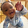 Jamila is registered to the contest to win money with this photo: baby, child, smiling, happy, curly_hair, onesie, blue_clothing, crib, wooden_crib, hand, face, crawling, indoor, mattress, patterned_mattress, closeup, portrait, infant, cute, playful